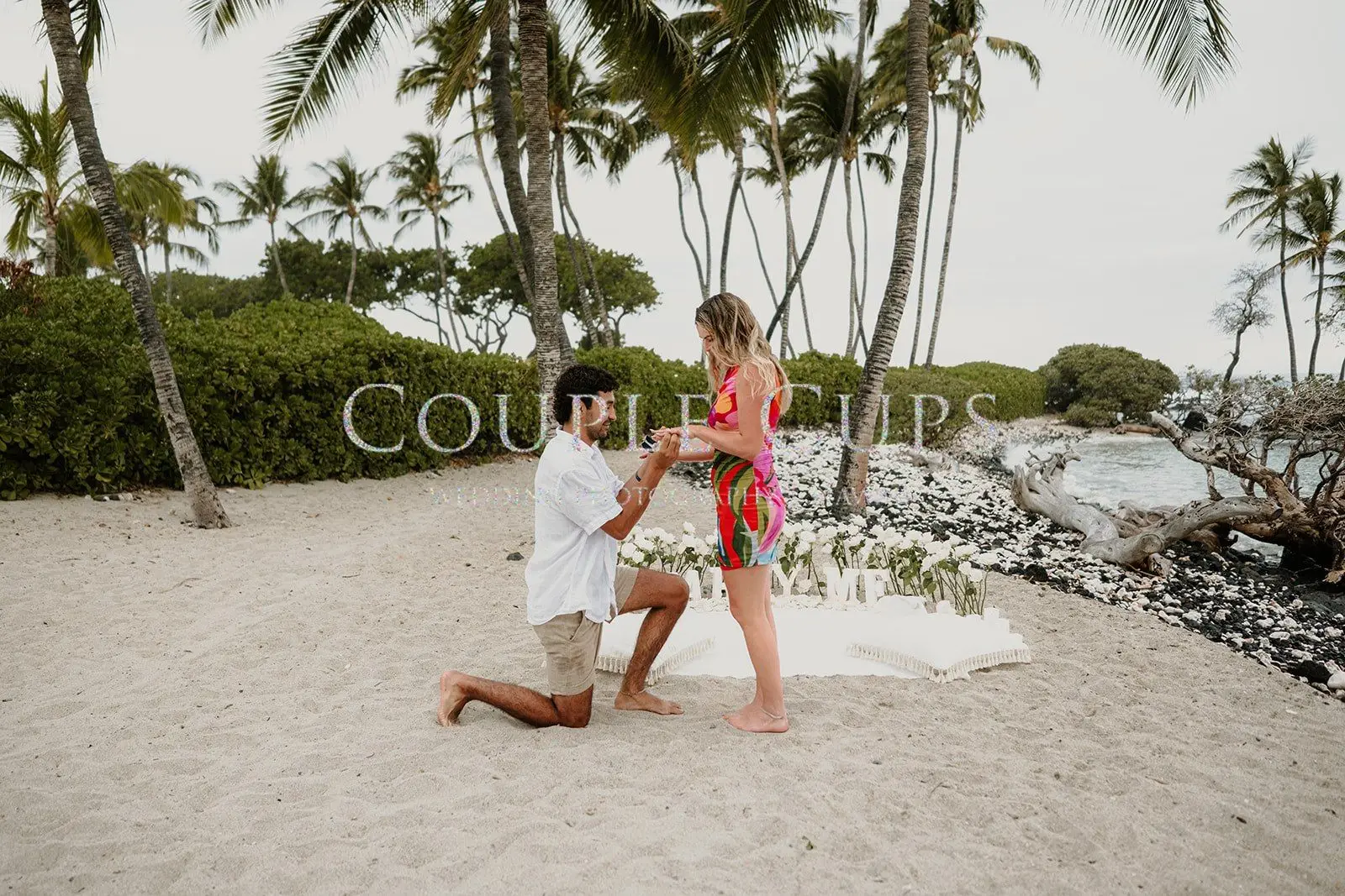 A couple embraces beneath tropical trees beside a flower-filled proposal setup
