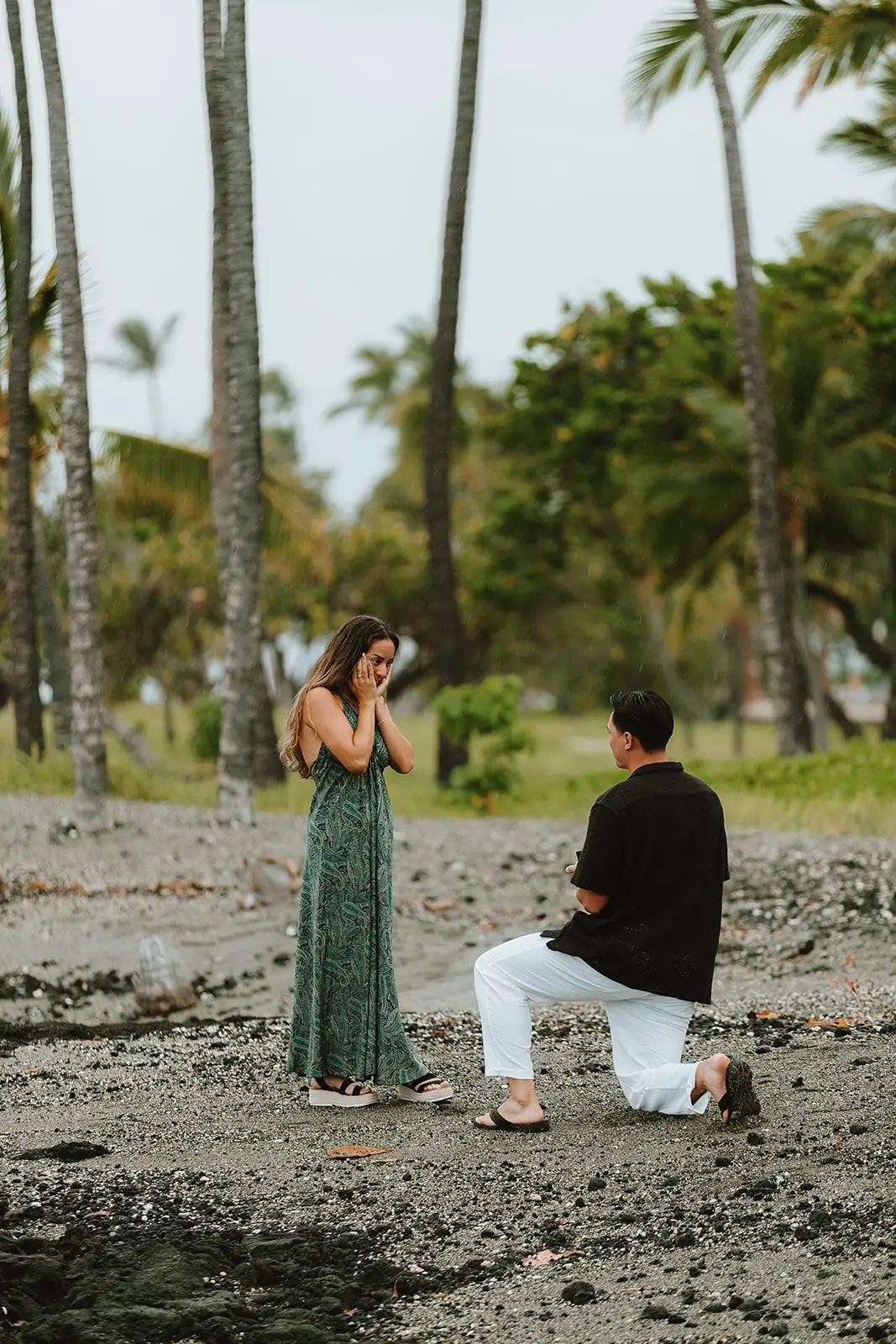 A proposal on black sand as one partner kneels and the other reacts in surprise